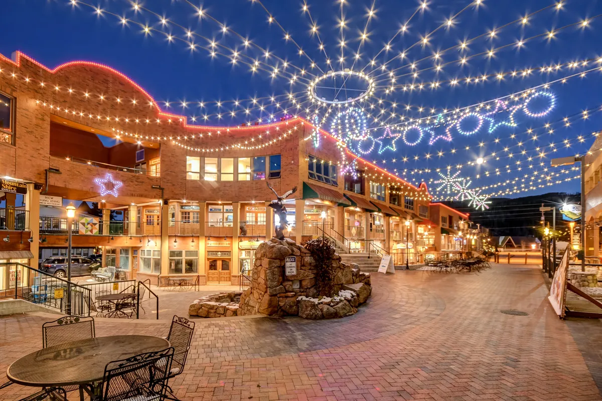 A lively evening exterior scene featuring twinkling string lights, decorative stars, an outdoor seating area, and a stone sculpture. The ambiance is festive and communal, perfect for evening strolls, dining, and participating in local events.