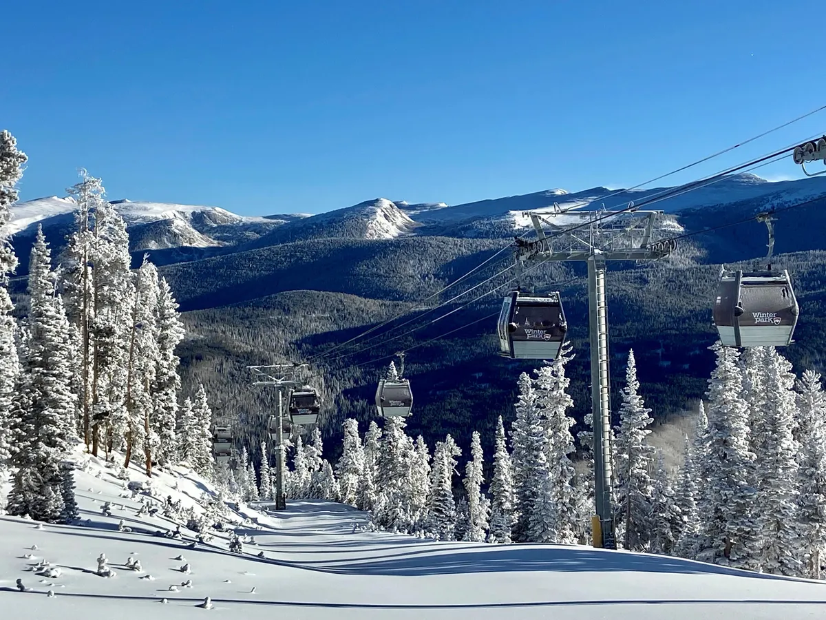 A dramatic, snow-covered landscape with a ski lift gliding above pine trees and mountains in the background. The scene captures the thrill and accessibility of winter sports, with skiers and snowboarders enjoying the pristine runs.