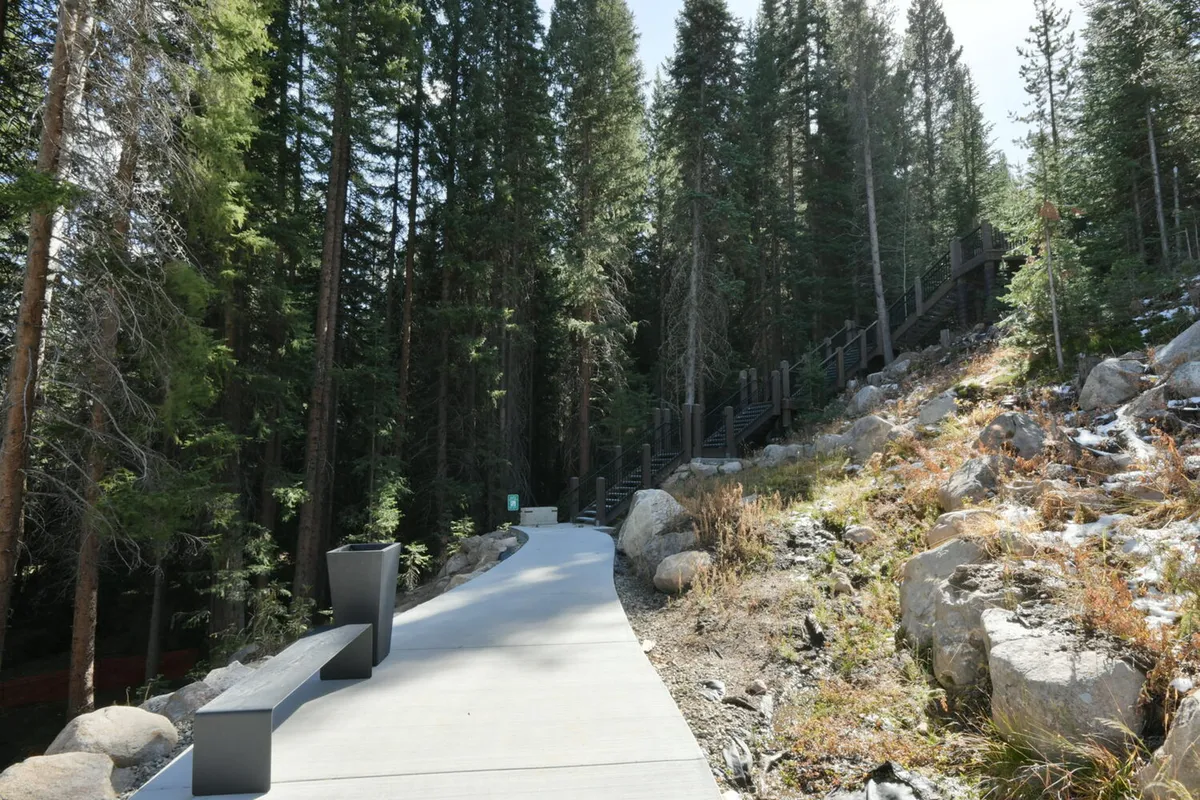 A serene exterior scene featuring a paved walkway winding among natural rock formations, tall trees, and a bench. The setting is lush and tranquil, inviting visitors to enjoy hiking or simply bask in the beauty of nature throughout the changing seasons.