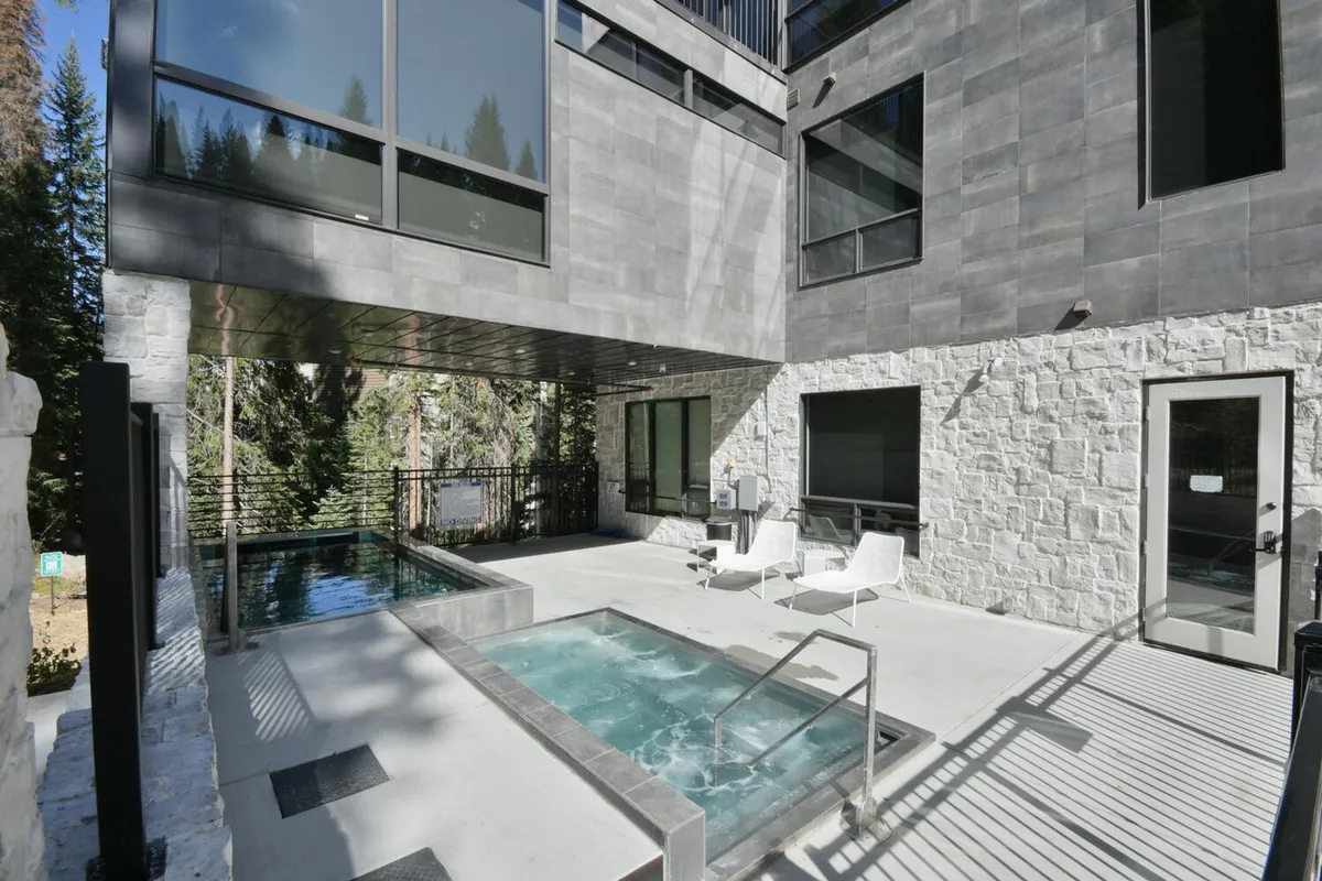 A luxurious outdoor lounge area featuring a sparkling hot tub and a swimming pool against a backdrop of modern architecture and wooded landscape. The space is designed for relaxation, social gatherings, and spa-like experiences, suitable for both day and night enjoyment.