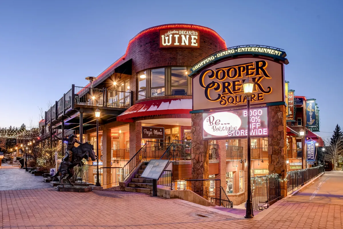 A lively shot of Cooper Creek Square's entrance, featuring the square's sign, decorative lighting, a wine shop, and a horse statue. People are mingling and moving about, suggesting a vibrant environment perfect for shopping, dining, and entertainment.
