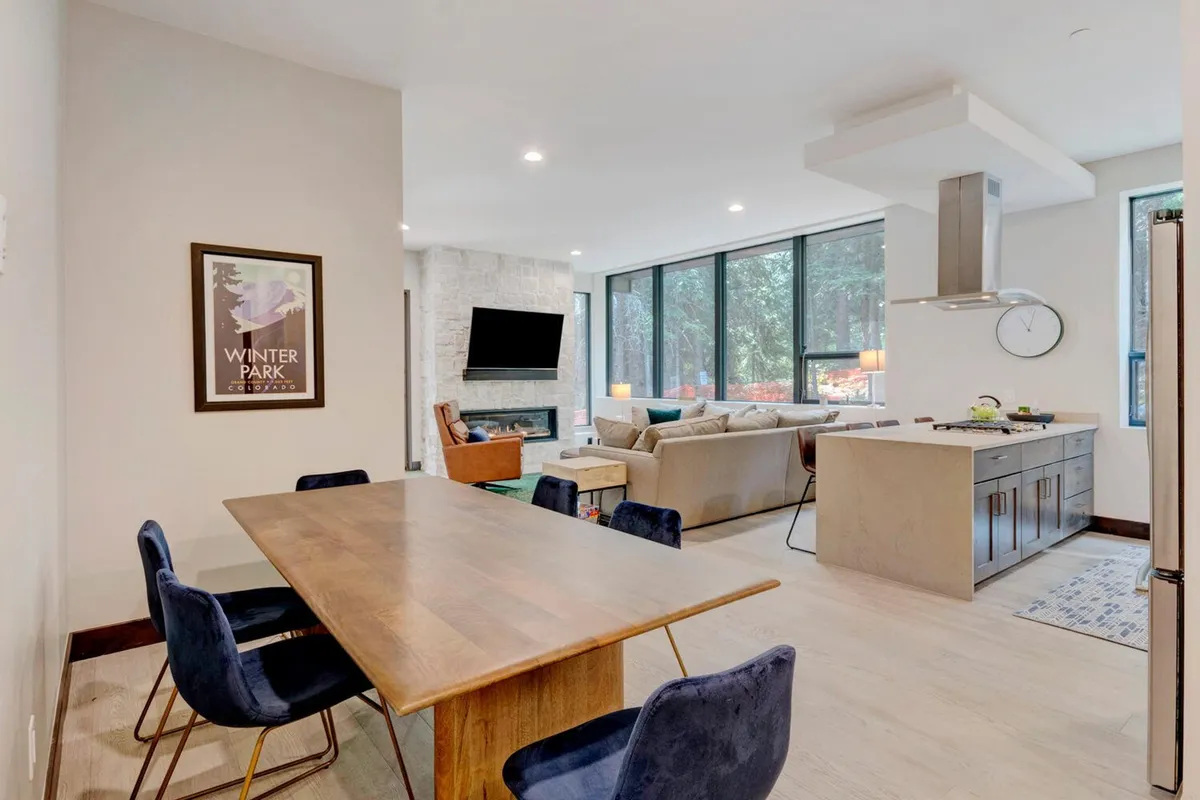 A wide-angle view of an open-concept living, dining, and kitchen space, featuring a cozy fireplace, a large dining table, a sleek kitchen island, and expansive windows revealing mountain scenery. The space is arranged for both socializing and relaxation, with plush seating and contemporary design details.