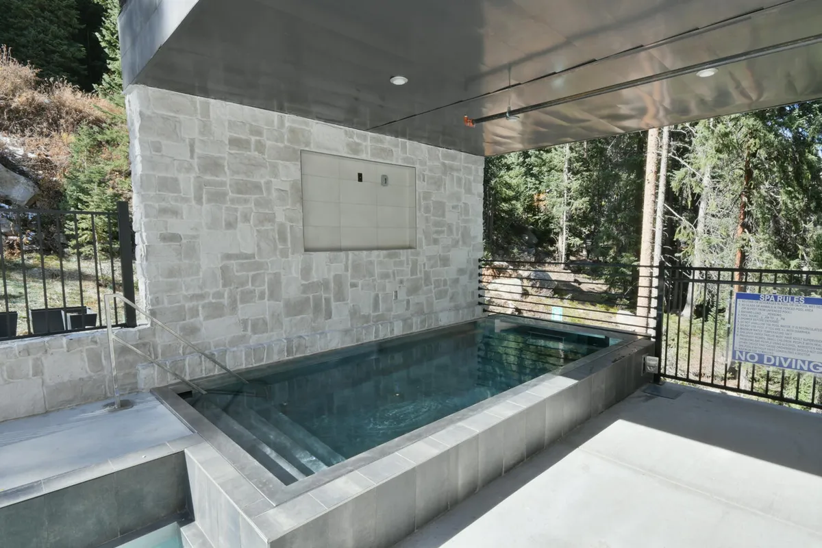 This exterior image features a serene hot tub area bordered by a rustic stone wall, with mature trees and a wooden fence providing a sense of privacy and immersion in nature. The setting suggests relaxation and socializing opportunities amid the beautiful Colorado landscape, with hints of mountain air and tranquil surroundings.