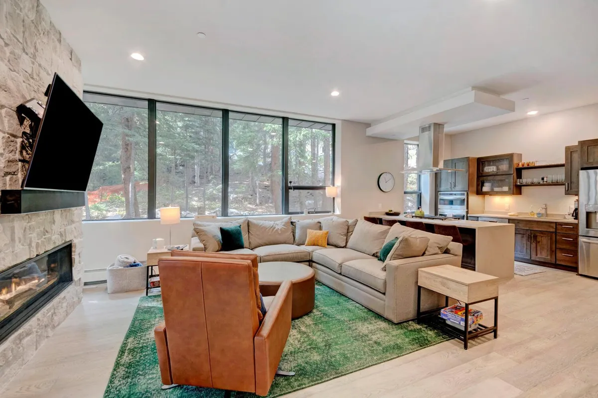 The image features a stylish, modern living area with a large sectional sofa centered around a warm fireplace, an open kitchen ideal for gatherings, and expansive windows that flood the space with natural light and mountain views. The space exudes comfort, perfect for relaxing after a day on the slopes or sharing meals with friends and family.