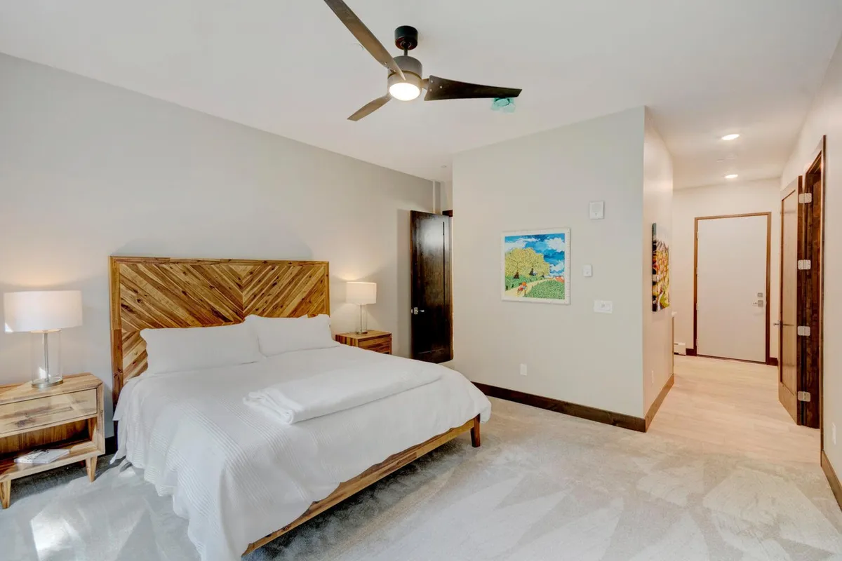 A serene bedroom featuring a bed with a wooden headboard flanked by table lamps and accented with artwork on the wall. Soft lighting and natural light streaming through the window create a tranquil, restful atmosphere.