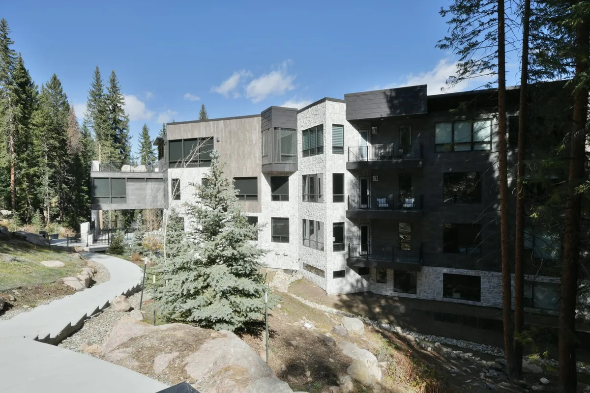 This exterior shot captures the property's stunning modern architecture, natural stone facade, and surrounding forest. A pathway leads up to the building, hinting at easy access to outdoor adventures and tranquil views from balconies or patios.