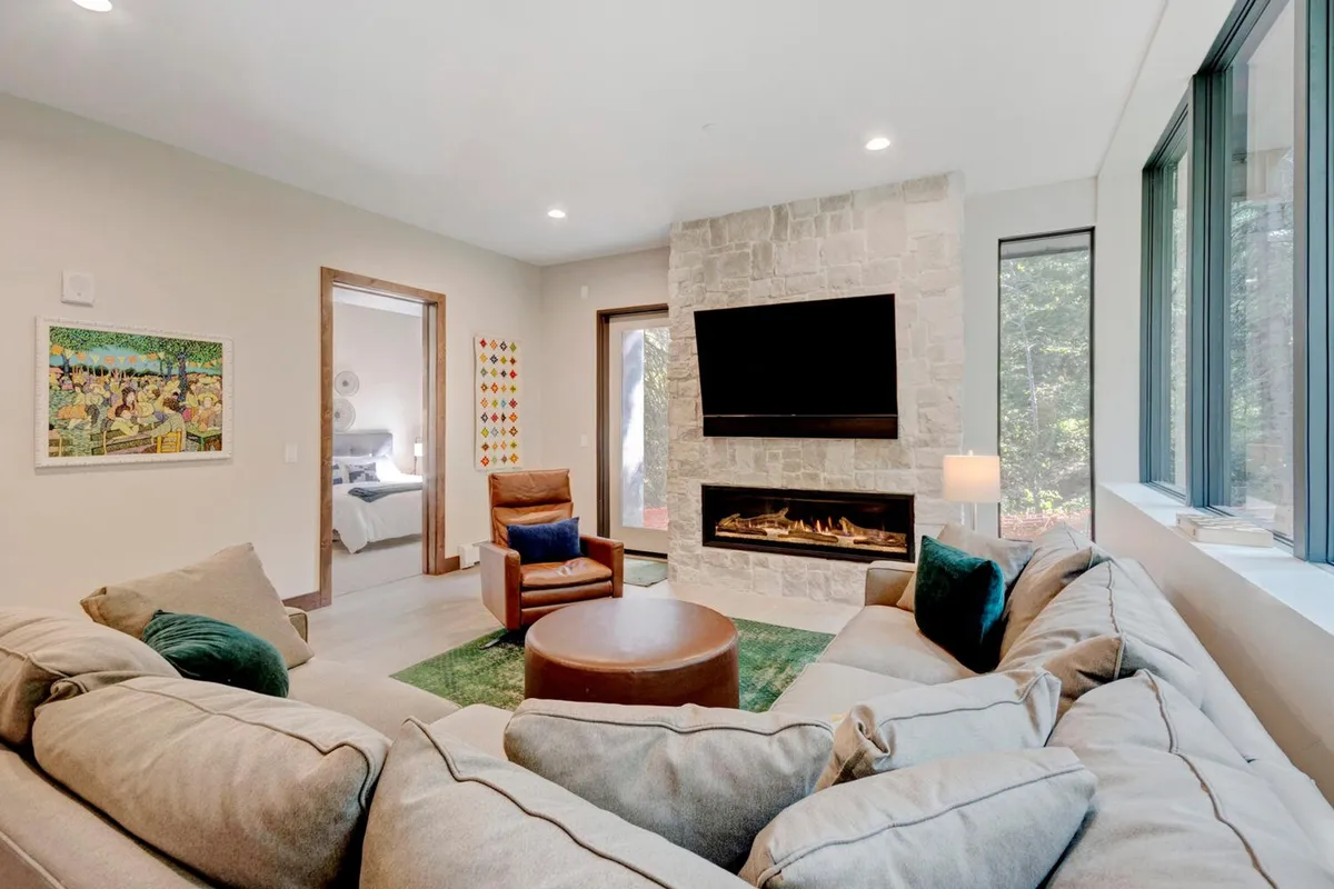A cozy living room anchored by a stone fireplace and plush sectional sofa, with a round coffee table and large windows framing peaceful views. The space is designed for relaxation and social activities like board games or movie nights.